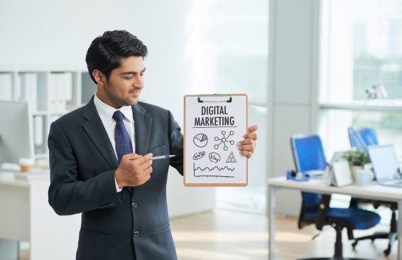 A businessman in a formal suit holding a clipboard labeled "Digital Marketing," pointing at marketing icons and graphs in an office setting.